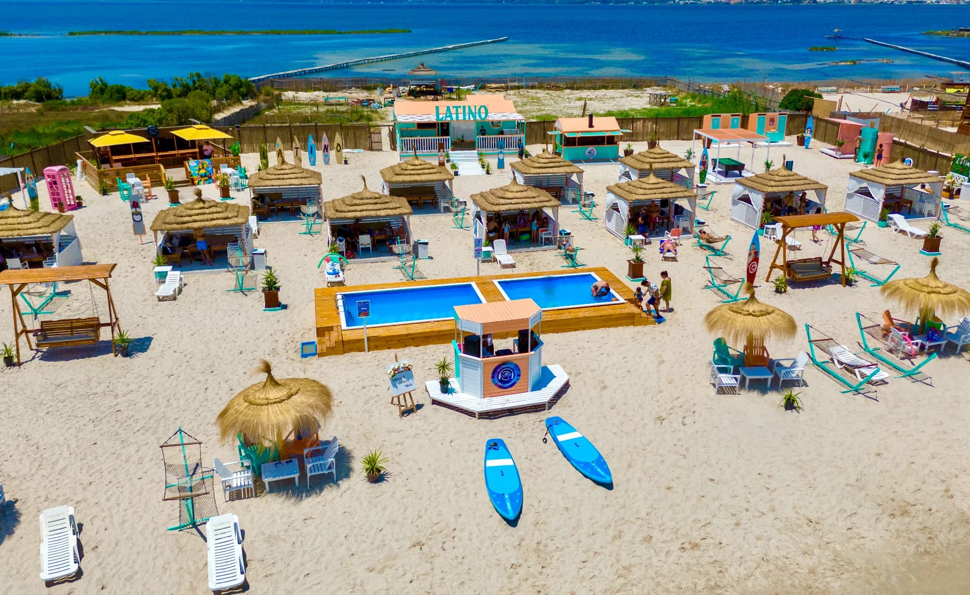 Une vue aérienne d'une plage avec une piscine, des cabines à toits de brise-soleil, des parasols et des chaises longues sur le sable.