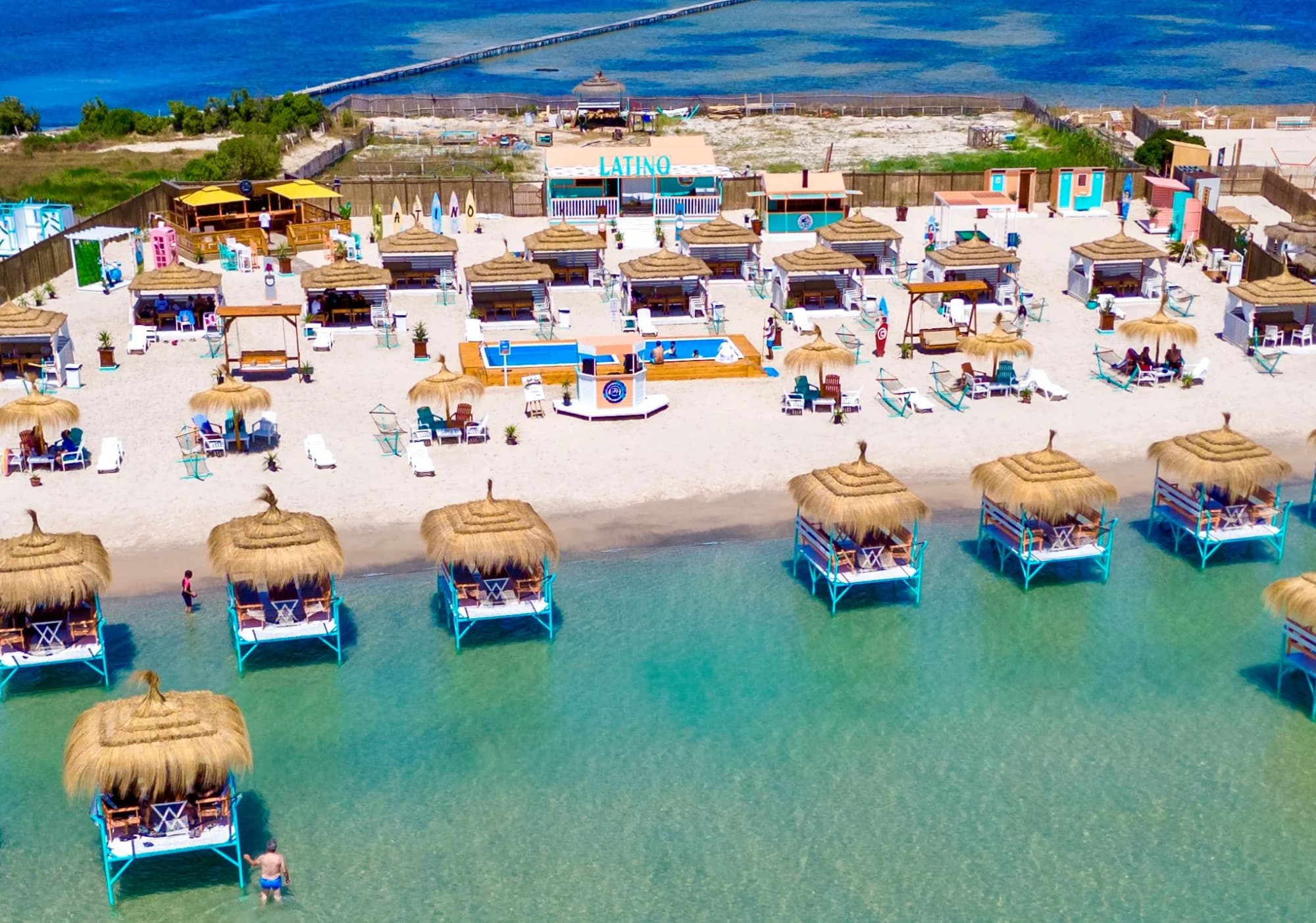 une plage avec des cabanas sur pilotis avec des toits en paille, des chaises longues colorées, et de l'eau turquoise, sous un ciel bleu clair, à latino coucou beach.